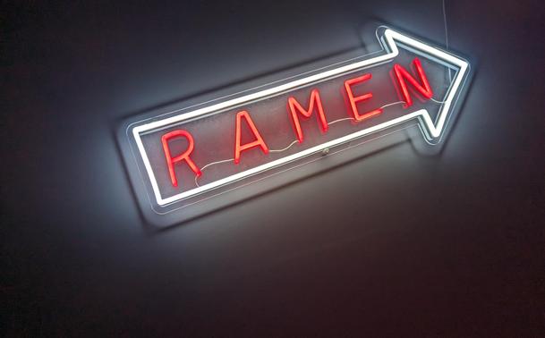 The image shows a close-up, slightly angled view of a neon sign. The sign is in the shape of a right-pointing arrow, outlined in a bright white neon tube. The word "RAMEN" is spelled out in bold, red neon letters within the arrow shape. The background is a dark wall, providing a stark contrast to the bright neon. The lighting is focused on the sign, highlighting its glow and the red lettering. The overall composition is simple and direct, drawing attention to the sign itself.