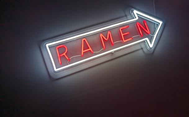The image shows a close-up, slightly angled view of a neon sign. The sign is in the shape of a right-pointing arrow, outlined in a bright white neon tube. The word "RAMEN" is spelled out in bold, red neon letters within the arrow shape. The background is a dark wall, providing a stark contrast to the bright neon. The lighting is focused on the sign, highlighting its glow and the red lettering. The overall composition is simple and direct, drawing attention to the sign itself.