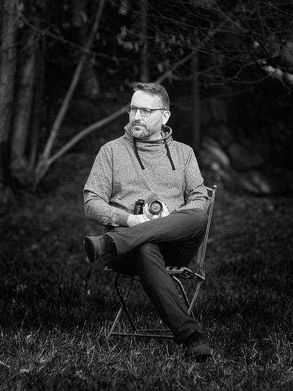 Male sitting on an iron chair in a garden, holding a camera with a brass lens in his lap.