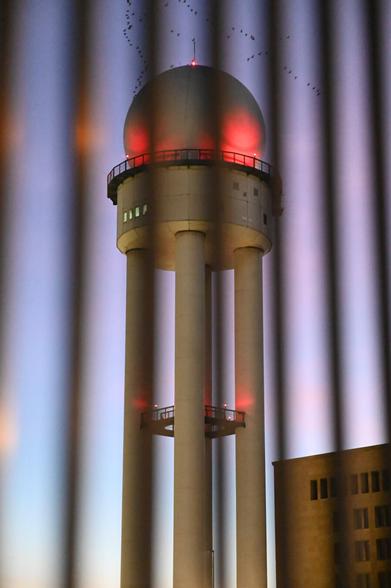 Ein hoher Turm mit einer großen Kuppel, beleuchtet von roten Lichtern, steht vor einem farbenfrohen Abendhimmel. Im Vordergrund sind vertikale Streifen zu sehen, die an ein Gitter erinnern. Eine Gruppe Vögel fliegt im Hintergrund.
A tall tower with a large dome, illuminated by red lights, stands against a colorful evening sky. Vertical stripes in the foreground resemble a fence. A flock of birds is flying in the background
Its located at the edge of tempelhofer feld which used to be an airport and was highly important for the berliners when the russians blocked Berlin and the airlift was installed.