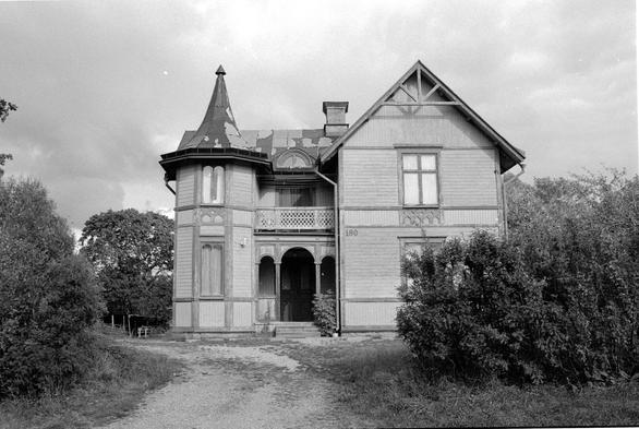 A black and white photograph of a old-style wooden house with a tin roof. It is summer and the bushes and trees around the house are lush.