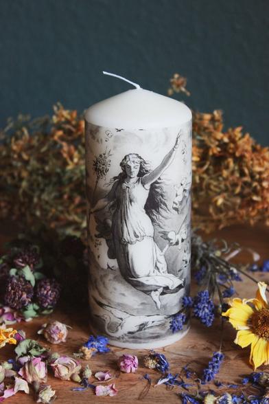 Das Bild zeigt eine fast weiße zylindrische Kerze mit einer detaillierten schwarz-weißen Illustration. Die Illustration zeigt eine Frau in einem fließenden Kleid, die einen Ast hält, umgeben von Vögeln und Wolken im Hintergrund.