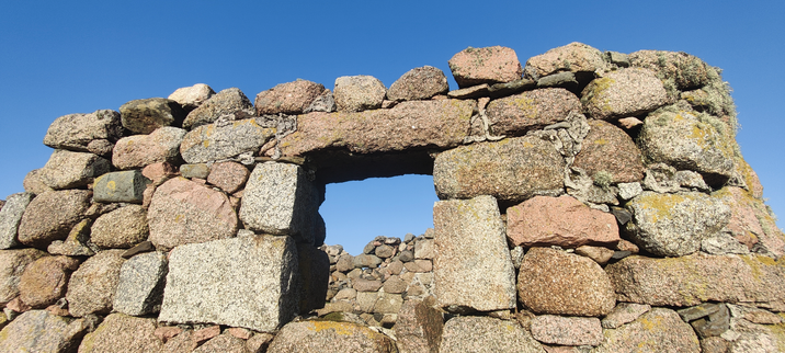 Looking in through the window in a ruined house wall made of natural, and roughly cut pink granite blocks. Behind, the inner house wall, and a blue blue sky above can be seen.