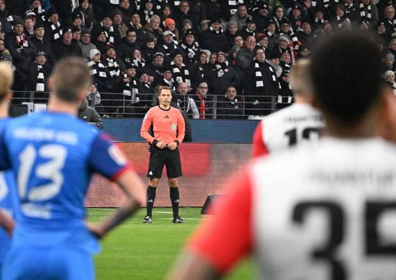 Ein Fußballschiedsrichter in einem pinkfarbenen Trikot steht auf dem Spielfeld, umgeben von Spielern beider Mannschaften, die im Vordergrund unscharf zu sehen sind. Im Hintergrund füllt eine große Menschenmenge das Stadion, viele Fans tragen schwarz-weiße Schals.