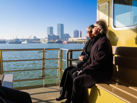 An eye-level shot shows two people sitting on a yellow bench on a ferry. The woman on the left is wearing sunglasses and a black coat. She has a black leather purse on her lap. The man on the right is also wearing sunglasses and a dark brown coat. In the background, there is a body of water with some ships, cranes, and buildings. The sky is blue and clear.