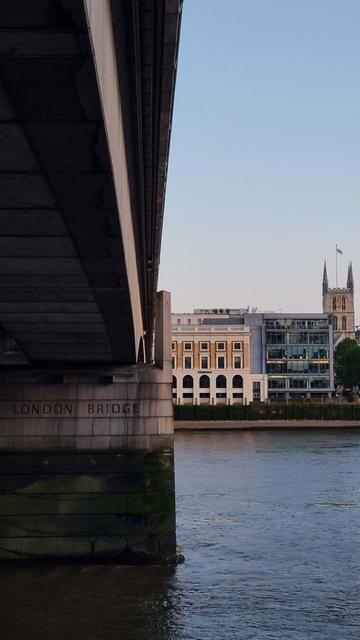 Picture from the underneath of London Bridge across the Thames. On the left in the bridge with pillar in the water and carving "London Bridge". On the right across the river are different buildings.