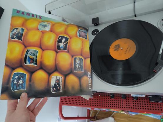 record player playing Bambix Leitmotiv LP, on the left side is a hand holding the cover. on the cover is a corn cob, on the corns are pictures