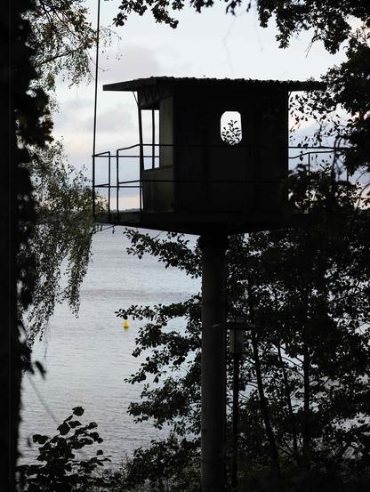 The image shows a silhouetted structure, possibly a small elevated lookout or guard tower, surrounded by trees. It's positioned near a body of water, visible in the background. The structure has a small window, and the scene is set against a cloudy sky.