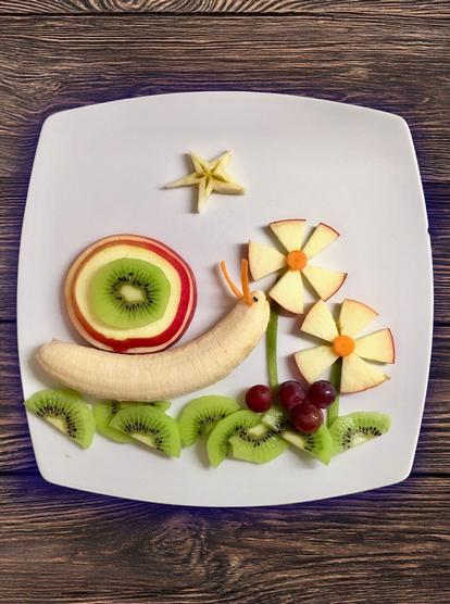 Ein Teller mit Obst. Das Obst zu einem Bild arrangiert. Eine Schnecke aus Banane mit einem Haus gestapelt aus Apfel- und Kiwi-Scheiben knabbert an einer Blume aus Apfelstückchen. Die  Mitte der Blume ist eine Karottenscheibe. Schräg darunter ist noch eine zweite Blume. Dunkle Weintrauben liegen unter ihnen. Der Boden ist aus halben Kiwischeiben gelegt.