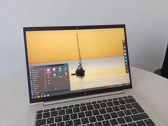 Ein silberner Laptop steht auf einem weißen Tisch. Auf dem Bildschirm des Laptops ist ein Hintergrundbild mit einem Segelboot auf dem Meer bei Sonnenuntergang zu sehen. Auf dem Bildschirm befindet sich außerdem ein Fenster mit einem Menü, das verschiedene Anwendungen auflistet.