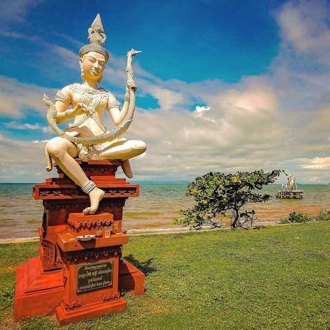 Photo shows a white statue of an Apsara lady demigod playing a Pin, an arched harp instrument. She is placed on a red fundament standing on a strip of green turf. In the background there is a a beach and sea stretching out. It is a sunny day, and most of the sky is deep blue. But in the horizon there are some light clouds showing.