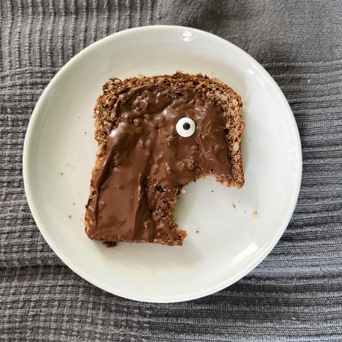 Angebissenes Brot mit Schokocreme. Durch den Biss rechts unten und ein Zuckerauge auf dem Brot entsteht der Eindruck eines schreienden Gesichts.