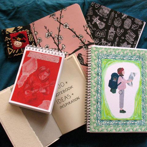 Pile of journals, some open, others closed. One has a sketch on red paper of a guitarist. Another has an image of a person with a backpack and map with washi tape around the border. Another says "bujo + notebook ideas + inspration".