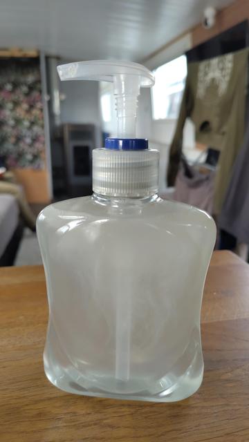 A close up of a soap dispenser, filled with soap from a refill shop, against a background of a widebeam canal boat interior with washing and a pellet stove visible.
