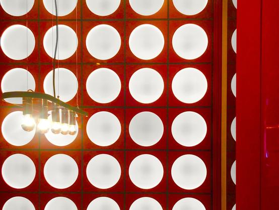Detail of a stylized wall in a small room. The wall is red with rows of large circular white lights evenly distributed across it. In the center-left of the photo  is a hanging lamp with  several clear bare light bulbs in a row. On the right of the frame is a vertical section of a solid red wall.
