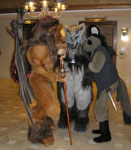 Three fursuiters - a manticore (Dar Rushk), a werewolf (Ysengrin), and a cat-raccoon (Kchierath) - in a tight group talking to each other, all facing inwards. They're in a hotel hallway with two elevators behind them.