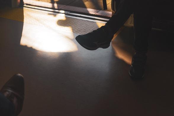The image shows a dimly lit interior scene, possibly in a vehicle or train, with partial views of two people. One person is sitting with their legs crossed, wearing dark pants and sneakers, while the shadows and sunlight create patterns on the floor. The setting suggests a moment of travel or commute.