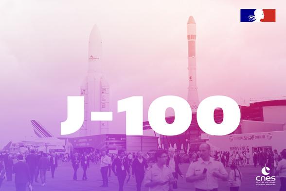 J-100 écrit en gros sur une photo prise au salon du Bourget 2023 avec Ariane 5 et Ariane 1 en fond sous un dégradé mauve