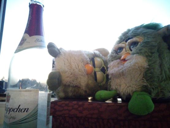 Eine leere Buddel alkfreier Ossisekt links. Zwei grüne Furbys vor dem Himmel rechts davon auf einer rot-lila Kiste. Der linke Furby liegt auf der Seite mit geschlossenen Augen. Der rechts hat die Augen halb zu und etwas den Schnabel offen.