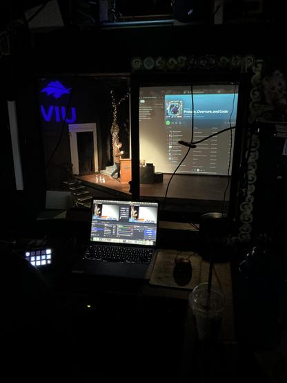 A dark photo from inside a control booth shows a laptop computer with OBS video control displays. Out the window is a theatre stage with a blue VIU logo on the black wall to the left.