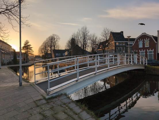 The sun is setting over a canal with a white bridge, the light reflecting on the water. Some trees in the background against the glowing sky. On the side of the canal you can see some Dutch houses