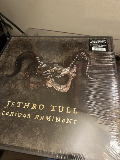LP, Jethro Tull, Curious Ruminant, still in shrink wrap with hype sticker top right corner. The main colour is brown and the primary image is a horned, scaly mask. Band name and LP title is in yellow under the mask.