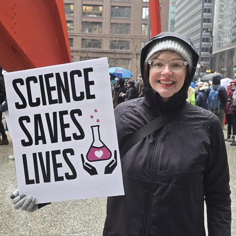 Woman in winter gear holds sign that says Science saves lives. Behind her is a crowd of protesters