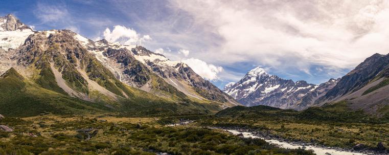 The image shows a panoramic view of a mountainous landscape with snow-capped peaks. The foreground features a grassy valley with a stream or river running through it. Clouds are present in the sky, adding to the scenic beauty of the area.