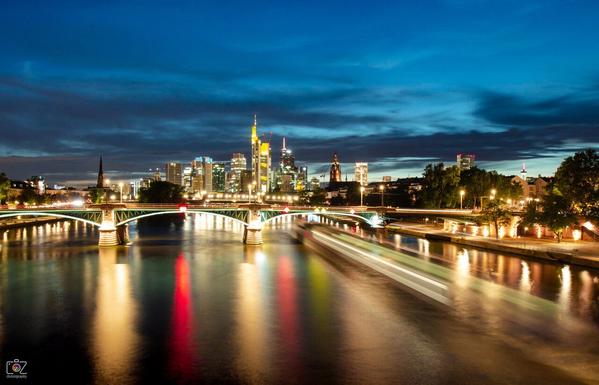 Skyline of the city of Frankfurt am Main during the blue hour at sunset in the evening. The river Main can be seen in the foreground, over which a road bridge with illuminated arches spans horizontally. Behind it in a bit of the distance is the skyline with illuminated skyscrapers. A few colorful lights are reflected in the water, and the light trail of a moving ship can be seen on the right edge (long exposure effect). The sky is slightly cloudy and blue.