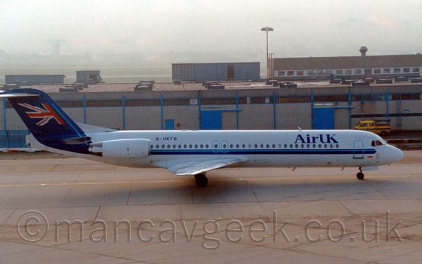 Side view of a white, twin engined jet airliner with the engines mounted on the sides of the rear fuselage.
There is a blue stripe running along the body, just under the passenger cabin windows, , getting wider as it sweeps up into the tail, which is filled with a waving half of the flag of the UK.
There are large blue "Air UK" titles on the upper forward fuselage.
Large concrete slabs of taxiway fill the foreground, with  sandy brown building with blue vertical support beams stretching across the background, with the airfield beyond being swallowed up in mist.