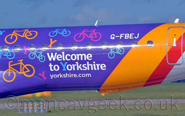 Close up of the purple rear fuselage of a jet airliner parked facing to the left.
There is white text "Welcome To Yorkshire" in the middle of the frame, surrounded by red, yellow, and blue images of bicycles and capital letter "Y"s, all seemingly at random angles.
The registration "G-FBEJ" is on the upper rear fuselage, on the right of the frame.
There are yellow, red, and white bands wrapped around the rear fuselage.
Green grass can be seen in the distance under the plane, with blue sky with hints of grey fluff filling the rest of the frame.