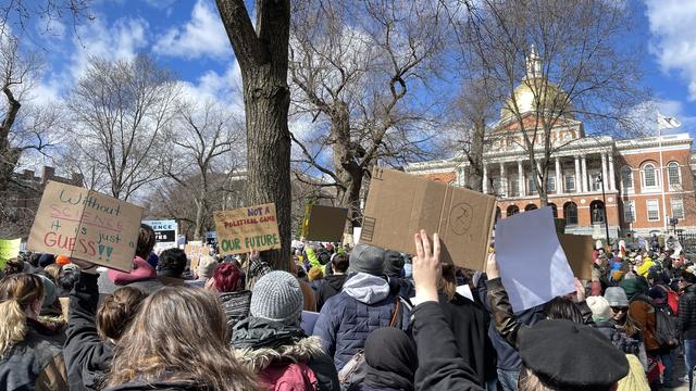 A crowd of several thousand people standing on a sloped lawn of the Boston Common, facing away from the camera toward the rally stage. Many in the crowd are holding up homemade signs which have clever science-related puns on them, or other science-themed messages. One says: "Without SCIENCE it is just a GUESS!!". Another says: "SCIENCE IS NOT A POLITICAL GAME It is the foundation of OUR FUTURE". In the background is the Massachusetts State House with its iconic gold dome. Above is a blue sky with a few fluffy clouds racing by on this windy day.