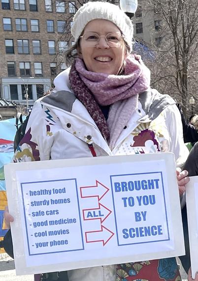 60-year-old white lady (me) bundled up against the cold and wind in a snowboard jacket, fleece scarf and knitted hat. I'm smiling broadly and holding a homemade sign that says: "Healthy food, sturdy homes, safe cars, good medicine, cool movies, your phone ---> ALL BROUGHT TO YOU BY SCIENCE"