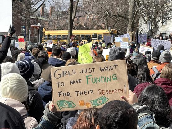 Someone in the crowd holding up a handwritten cardboard sign that says "GIRLS JUST WANT TO HAVE FUNDING FOR THEIR RESEARCH"