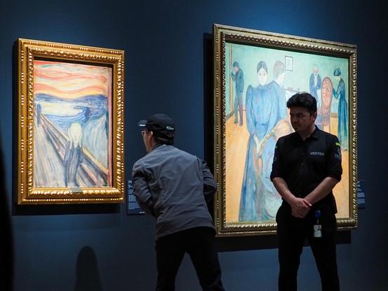 A person wearing a baseball hat looking intensely at Edvard Munch"s famous painting The Scream as close as it was allowed to. Next to him to the right is a security guard watching him like a hawk. Behind the security guard is another, larger painting of Munch's.