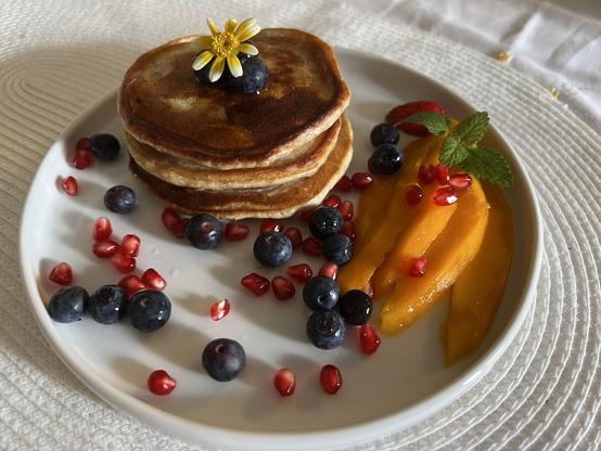 Bild eines Stapels aus drei Pancakes mit Heidelbeeren und Granatapfellernen, geschnittener Mango und etwas Minze und einer Blume als Deko auf einem weißen Teller.