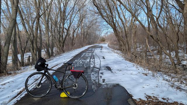 Dark brown Diverge bicycle with Bar Mitts parked on the asphalt Douglas State Trail looking North just past the trailhead. The trail is wet and partially snow covered, and is surrounded by bare trees. The sky is blue.