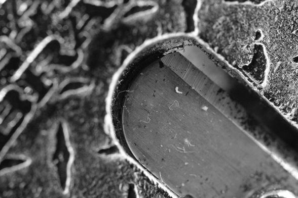 The image is a close-up, black-and-white photograph of a textured surface. The main focus is on what appears to be the tread of a shoe or tire, with a distinct pattern of grooves and ridges. The tread pattern is intricate, with deep channels that are likely designed for grip and traction. The surface of the tread shows signs of wear and has small particles or debris embedded in it, suggesting it has been used. The background is out of focus, emphasizing the texture and details of the tread. The overall composition highlights the contrast between the smooth and rough areas of the tread.