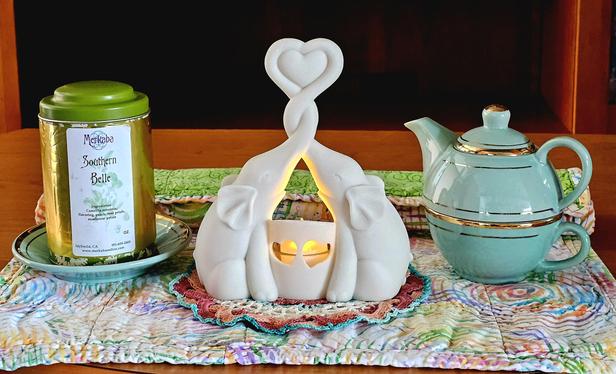 Tea tableau with a bisque candleholder in the shape of a pair of elephants, trunks entwined to form a heart. They are surrounded by a mint green tea-for-one set with gold leaf trim and a green and gold tea tin of Southern Belle black and green tea blend flavored with peach, rose, and sunflower petals. They sit upon a quilt square patterned in pastel candy- colored spirals.