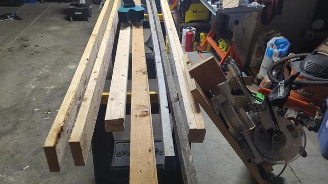 a garage containing two sawhorses, which are loaded up with eight 2x4 beams of various lengths and two 2x6 beams that are eight feet long. to the right, a chop saw rests on the floor leaning against the sawhorses. An oddly shaped metal bracket rests on some of the 2x4s, its purpose unclear.