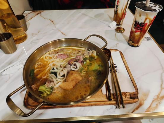A photo of a table of food. You see a large copper bowl filled with meat, noodles, and vegetables floating in a spicy looking broth. Two brown sugar boba teas are also visible.