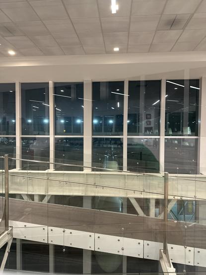 Interior view of a modern airport terminal with large glass windows and overpass walkways. The scene is illuminated with artificial lighting, showcasing a nighttime ambiance outside where aircraft are visible.