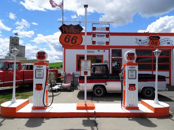 Vintage Phillips 66 Gas Station restored to bright colors of orange, black, and white. Two restored vintage gas pumps stand on a concrete island painted bright white with orange trim. Behind them, a cinder block building with orange and black signs that say "Classic Phillips 66, Steptoe, WA, Trop Artic Motor Oil, Mechanic on Duty, 24-hour towing service." Two vintage pickup trucks, one red and the other black, red, and silver. An American flag flies on the building's roof against a blue sky with fluffy white clouds.
