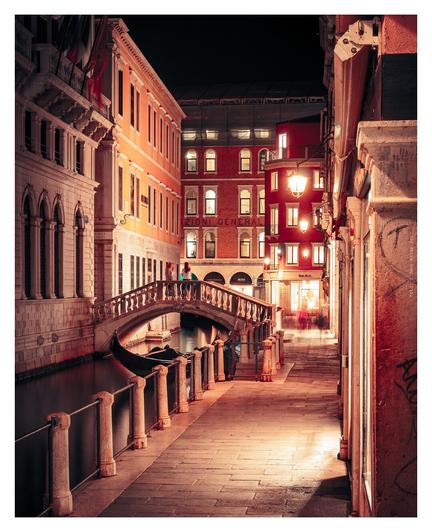 A serene night view of a bridge arching over a tranquil canal in Venice, illuminated by soft lights reflecting on the water.