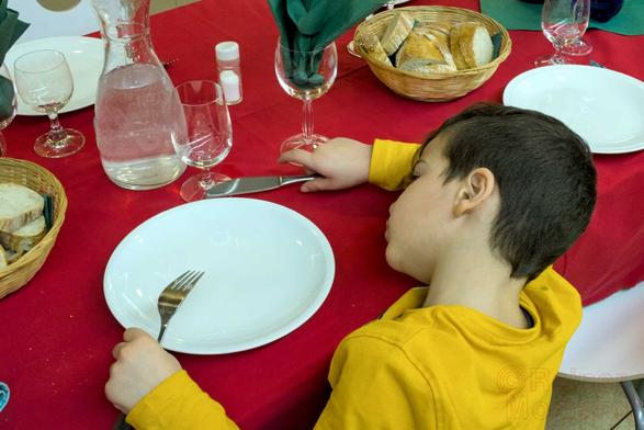 Farbfotografie im Format 3:2 waagerecht.
Auf einem Tisch, mit rotem Tischtuch, stehen Teller, Gläser, eine Wasserkaraffe, Brotkörbchen. Am Tischrand sitzt ein Kind mit gelbem Sweatshirt, Gabel inks und Messer rechts in der Hand, auf dem Tisch, neben einem weißen Teller. Der Kopf des Kindes ist, seitlich auf den rechten Oberarm gelegt.