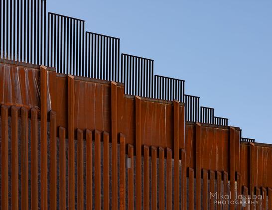 The rusty steel U.S.-Mexico border fence as an abstract photograph. In the front, the new fence is a series of bright, rusty square steel tubes with a 4' tall flat rusty steel plate welded across the tops, supposedly to prevent people from climbing over. Behind the new fence, up on a hill above it, is the Obama-era fence, shorter and without the no-climb plate on top. Like the new fence, it descends a slight slope in stair-step fashion.