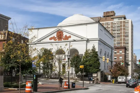 The image depicts a white building with a domed roof, featuring classical architectural elements such as large arches and decorative details. The structure appears to be situated in an urban environment, surrounded by trees and nearby city buildings. There are also construction cones visible in the foreground, indicating road work or other urban maintenance. The overall scene suggests a blend of historical and modern elements in the surrounding cityscape.

Image Credits: Wikimedia / Acroterion / CC BY-SA 4.0