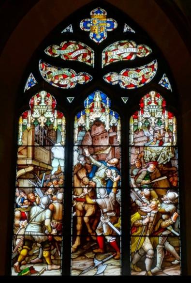 The top panel of the stained glass window of St. Giles. This depicts the Earl of Moray (in Knight's armour and a blue cape-cloak) being shot by a wide-barrelled gun (left). The crowd is shocked and afraid, many are looking up towards the killer, who is in a nearby balcony.