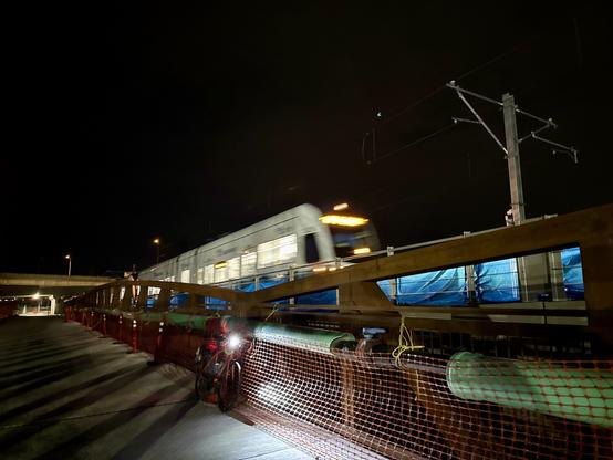 A light rail train passes by on an elevated bridge next to my bicycle resting on a trail bridge at night