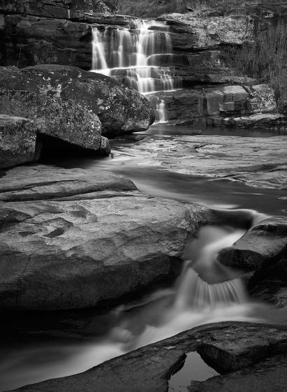 The photo shows a waterfall in the upper middle, where the flowing water looks milky due to the long exposure. The water is the running over rocky ground towards another small waterfall / step towards the front left. The photo is black and white.